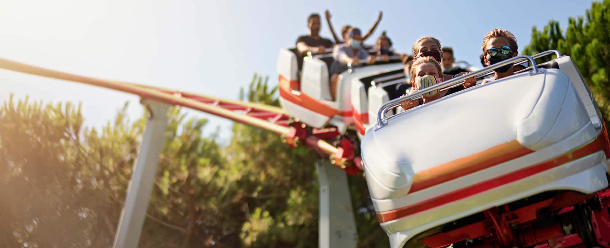 Three mask-wearing kids ride in the front car on a white and red roller coaster